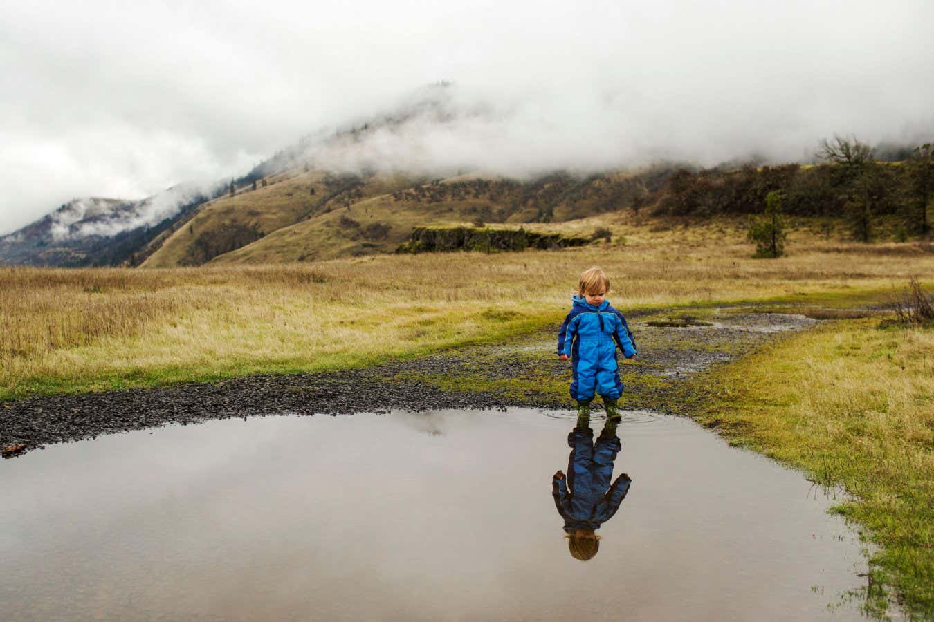 2HXRJ2C A young child stands at edge of calm pool with clouds over hills