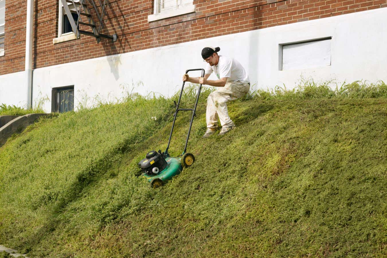Man mowing downhill Description Man wearing white overalls and back to front baseball cap trying to mow down steep verge outside building. Memphis, USA